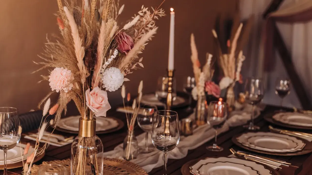 Stół weselny w stylu boho z bukietem z traw pampasowych, róż i świecami. Eleganckie nakrycie z złotymi talerzami i kieliszkami.