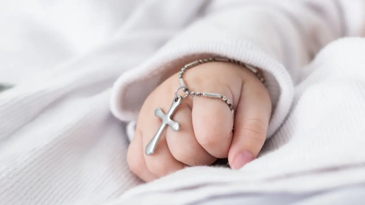 Dziecięca dłoń trzyma krzyżyk na łańcuszku, symbol tego, co jest potrzebne do chrztu.