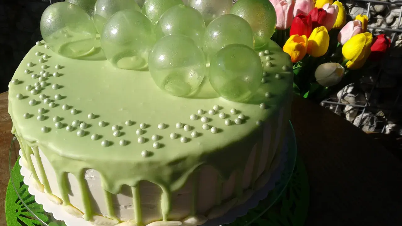 Zielony tort udekorowany koralikami i przezroczystymi, zielonymi balonami z żelatyny. Obok bukiet kolorowych tulipanów.