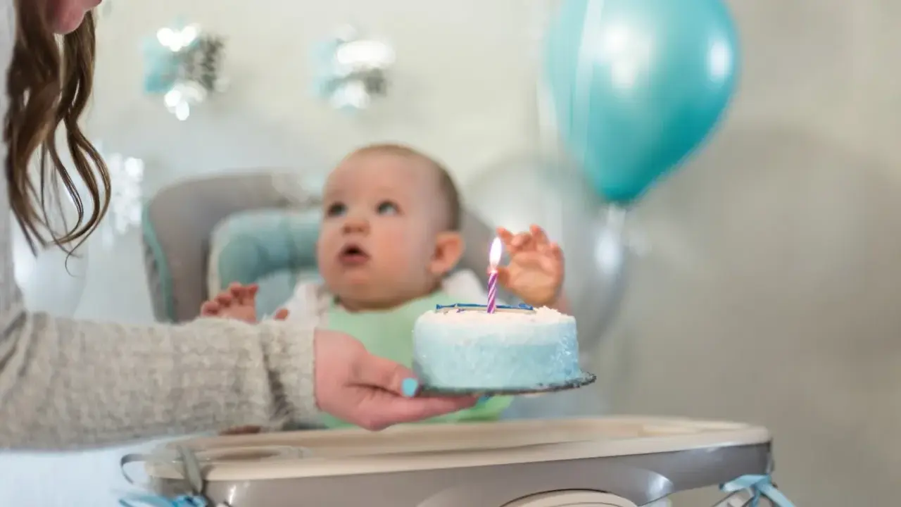 Mama podaje tort z jedną świeczką na roczek maluszkowi. Balony i dekoracje tworzą radosną atmosferę.