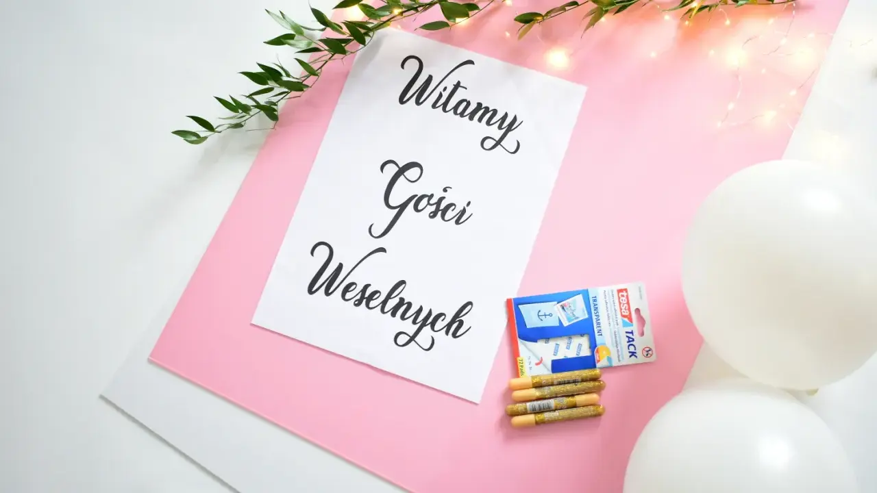 Tablica powitalna wesele z napisem "Witamy Gości Weselnych", ozdobiona gałązką zieleni i lampkami. Obok taśma, złote pisaki i białe balony.