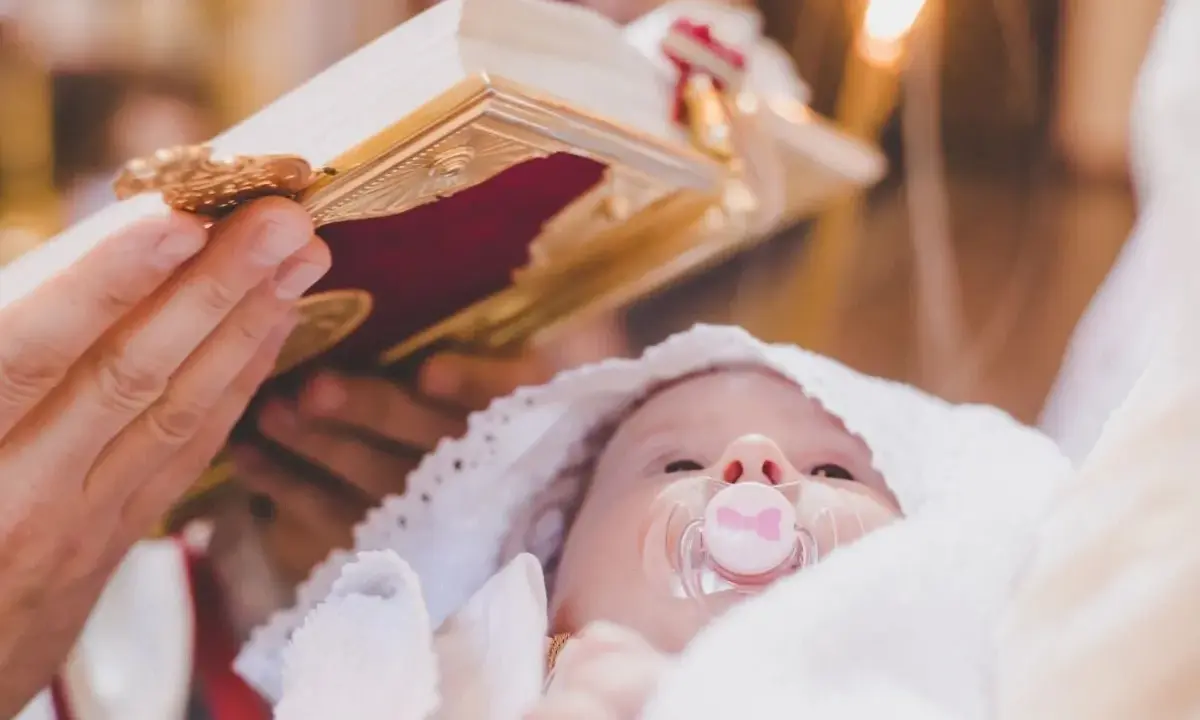 Dziecko z chupkiem w buzi, otulone białym materiałem, podczas ceremonii chrztu. Nad nim trzymana jest księga, a w tle płonie świeca.