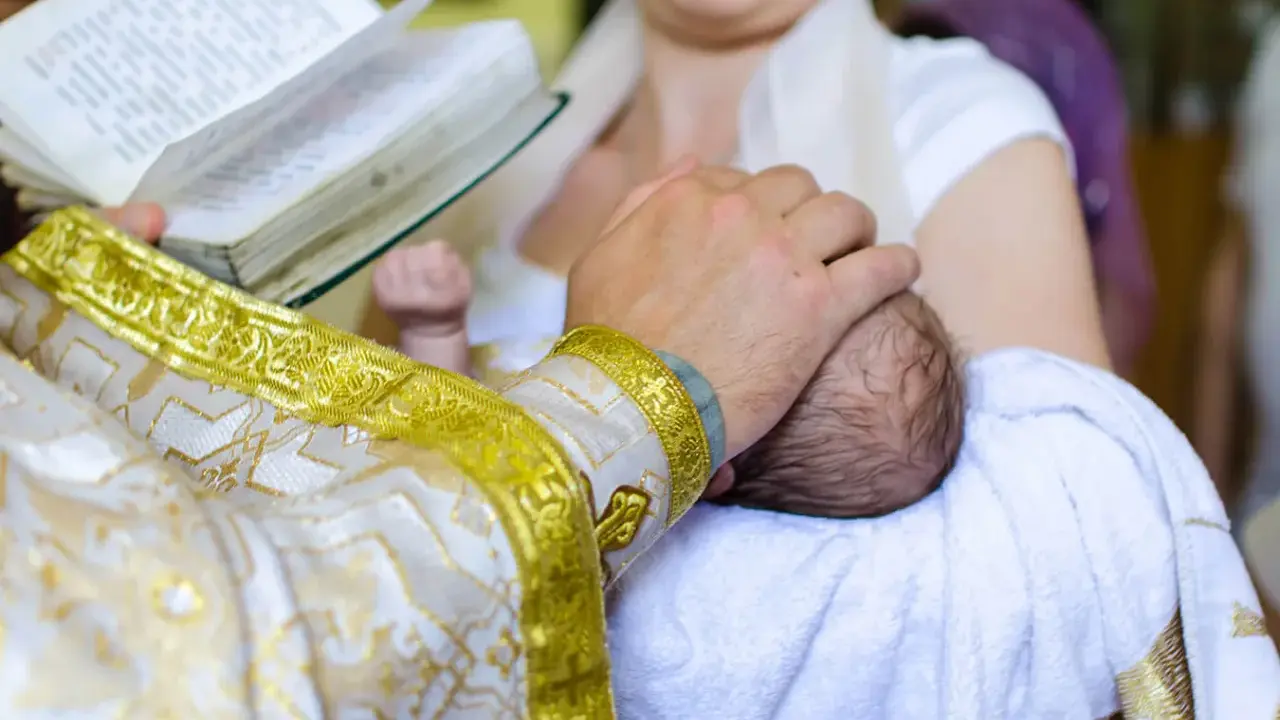 Ksiądz w złotej szacie trzyma dziecko do chrztu, czytając z księgi.