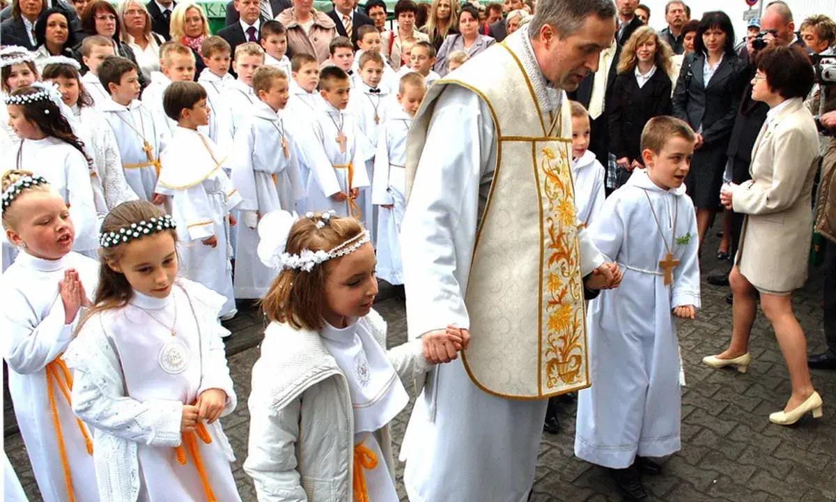 Dzieci w białych strojach, kiedy jest komunia, idą z księdzem.