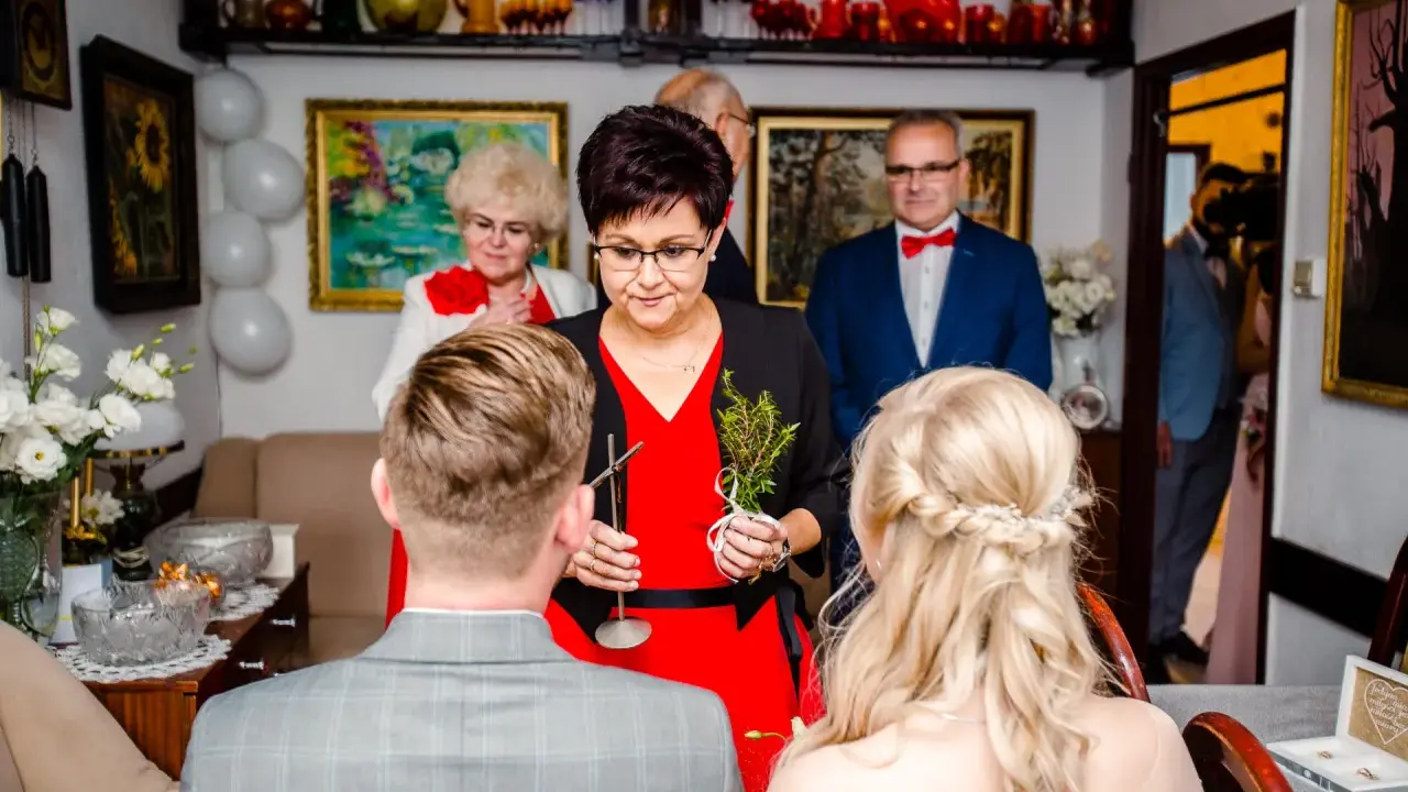 Rodzina udziela błogosławieństwa młodej parze. Kobieta trzyma gałązkę ziół, symbolizującą pomyślność.