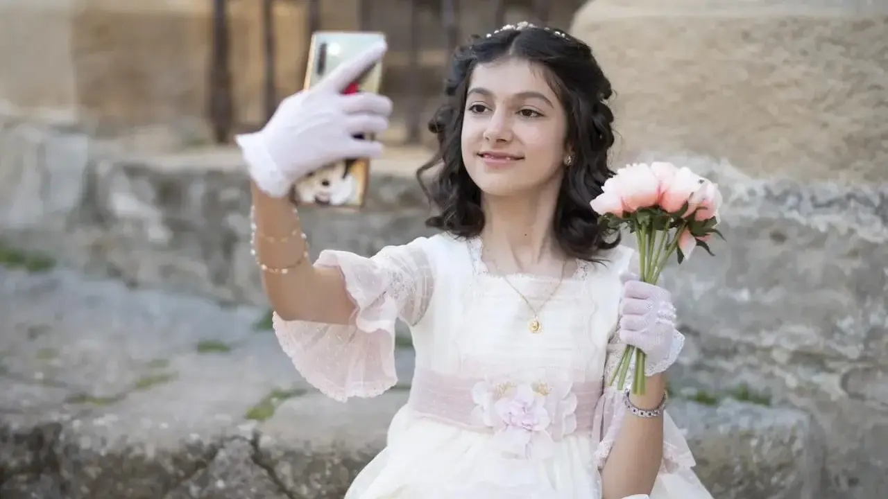 Dziewczynka w sukni komunijnej robi selfie z bukietem kwiatów. To ważny moment, przygotowania do pierwszej komunii świętej krok po kroku.