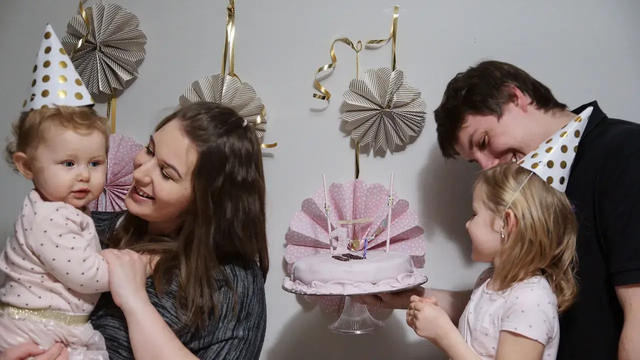 Rodzina świętuje roczek dziecka. Mama trzyma malucha, a tata podaje tort. W tle wiszą papierowe dekoracje, idealne, jak zrobić dekoracje na roczek.