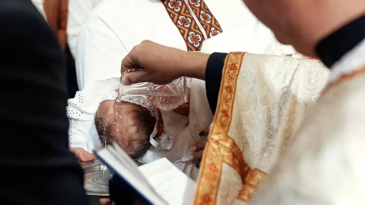 Chrzestni słuchają słów kapłana, gdy ten polewa wodą głowę niemowlęcia.