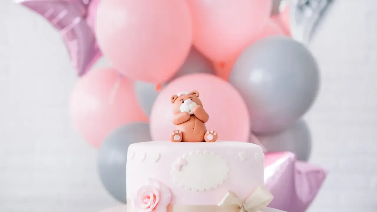 Uroczy topper na tort baby shower w kształcie misia siedzącego na różowym torcie, otoczony balonami w pastelowych kolorach.