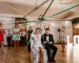 Panna młoda i pan młody siedzą pod baldachimem z kolorowych wstążek, co zamiast oczepin. Goście trzymają wstążki, tworząc magiczną atmosferę.