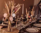 Stół weselny w stylu boho z bukietem z traw pampasowych, róż i świecami. Eleganckie nakrycie z złotymi talerzami i kieliszkami.