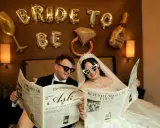 Para młoda i pan młody czytają gazetę "The Wedding Post". Dekoracje "Bride to Be" i balony w kształcie kieliszka i butelki szampana sugerują wieczór panieński lub kawalerski. Kiedy dać zawiadomienie o ślubie w pracy?