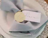 Elegancka winietka z imieniem "Peram" na jasnoszarym obrusie, ozdobiona okrągłym, żółtym tagiem.