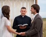 Ksiądz mówi na ślubie, trzymając otwartą księgę. Panna młoda i pan młody trzymają się za ręce, patrząc na siebie z uśmiechem.