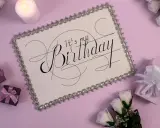 Eleganckie zaproszenie na urodziny w stylu savoir vivre. Biała kartka z ozdobnym napisem "It's my Birthday", otoczona prezentami i świecą.