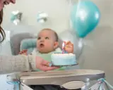 Mama podaje tort z jedną świeczką na roczek maluszkowi. Balony i dekoracje tworzą radosną atmosferę.
