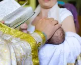 Ksiądz w złotej szacie trzyma dziecko do chrztu, czytając z księgi.