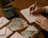 Ręka zapisuje w notesie, co zrobić z pieniędzmi z wesela. Obok leżą koperty, banknoty 100 zł i kalkulator.
