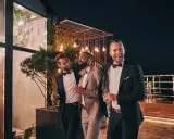 Trzech eleganckich panów, w tym drużba na weselu, śmieje się i trzyma drinki na tarasie nocą, wśród ozdobnych świateł.