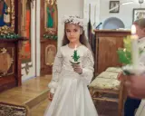 Dziewczynka w białej sukni i wianku trzyma zapaloną świecę. To ważny dzień, w którym klasa przystępuje do komunii.
