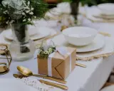 Elegancki prezent ślubny DIY, ozdobiony gałązkami i białymi kwiatami, czeka na stole obok złotych sztućców i świecznika.