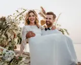 Szczęśliwa para świętuje ślub bez wesela. Pan młody niesie pannę młodą na rękach przy ozdobnym łuku.