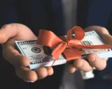 Dłonie trzymają plik banknotów 100-dolarowych przewiązany czerwoną wstążką. To pieniądze zamiast prezentów na urodziny, praktyczny i cenny podarunek.