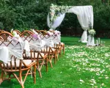 Eleganckie dekoracje na ślub cywilny w plenerze: rzędy krzeseł ozdobionych białymi wstęgami i kwiatami, łuk ślubny z tkaniną i zielenią, ścieżka usypana płatkami róż.