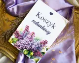 Eleganckie elementy zaproszenia: kartka z napisem "Koszyk ratunkowy", ozdobiona bukietem bzu, na tle fioletowej satyny i złotej ramki.