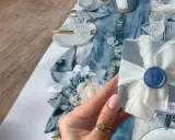 Elegancki stół na chrzciny: błękitne serwetki, złote sztućce, kwiaty i pudełeczko z odciskiem stópki.