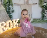 Urocza dziewczynka w różowej sukience świętuje pierwszy roczek. Litery "ROK" i kwiaty tworzą piękny motyw przewodni na jej wyjątkowy dzień.