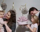 Rodzina świętuje roczek dziecka. Mama trzyma malucha, a tata podaje tort. W tle wiszą papierowe dekoracje, idealne, jak zrobić dekoracje na roczek.