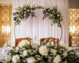Elegancki ołtarz na ślub w plenerze, ozdobiony białymi kwiatami i zielenią na złotym okręgu.