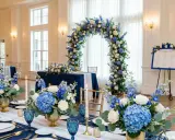 Elegancka sala weselna z niebieskim jako kolorem przewodnim. Dekoracje z niebieskich i białych kwiatów, złote wazony i świece tworzą romantyczną atmosferę.