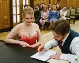 Panna młoda w czerwonej sukni i pan młody podpisują dokumenty. W tle goście robią zdjęcia telefonami. Może to być ślub, gdzie nie mam świadków.
