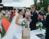 Para młoda podczas ceremonii ślubu cywilnego na zewnątrz. Panna młoda w sukni, pan młody w garniturze. Goście obserwują.