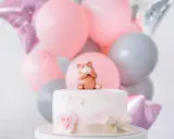 Uroczy topper na tort baby shower w kształcie misia siedzącego na różowym torcie, otoczony balonami w pastelowych kolorach.