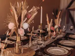 Stół weselny w stylu boho z bukietem z traw pampasowych, róż i świecami. Eleganckie nakrycie z złotymi talerzami i kieliszkami.