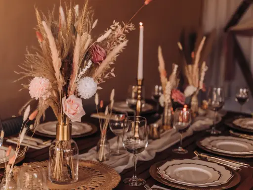 Stół weselny w stylu boho z bukietem z traw pampasowych, róż i świecami. Eleganckie nakrycie z złotymi talerzami i kieliszkami.
