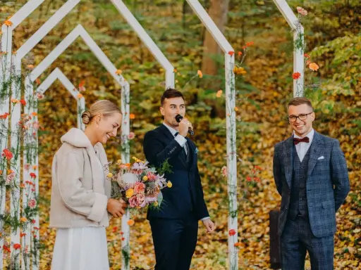 Piękna panna młoda z bukietem, pan młody przemawia do mikrofonu, a pan młody z uśmiechem. Wesele jesienią w otoczeniu kolorowych liści.