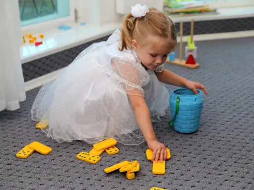 Mała dziewczynka w białej sukience bawi się klockami domino, tworząc kącik zabaw dla dzieci na weselu.