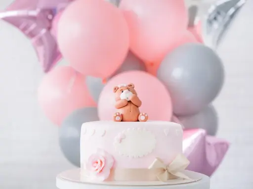 Uroczy topper na tort baby shower w kształcie misia siedzącego na różowym torcie, otoczony balonami w pastelowych kolorach.