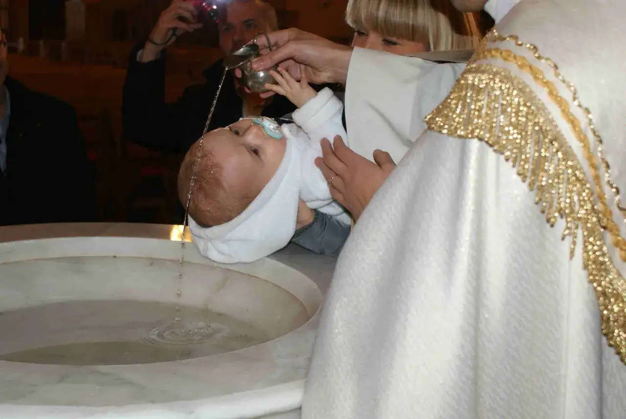 Kapłan polewa wodą głowę dziecka, wykonując formułę chrztu. W tle widać rodziców i fotografa.