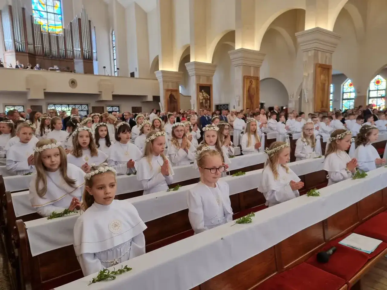 Dzieci w białych strojach, z wiankami na głowach, siedzą w ławkach kościoła. To ważny dzień, kiedy jest komunia.