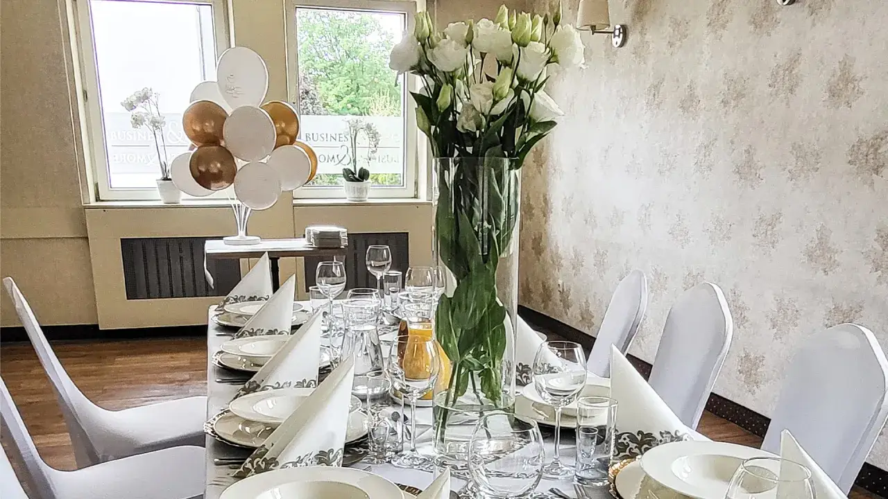 Elegancko nakryty stół w restauracji, ozdobiony białymi kwiatami i balonami. Idealne miejsce na uroczystość, jak chrzciny, które mogą trwać wiele godzin.
