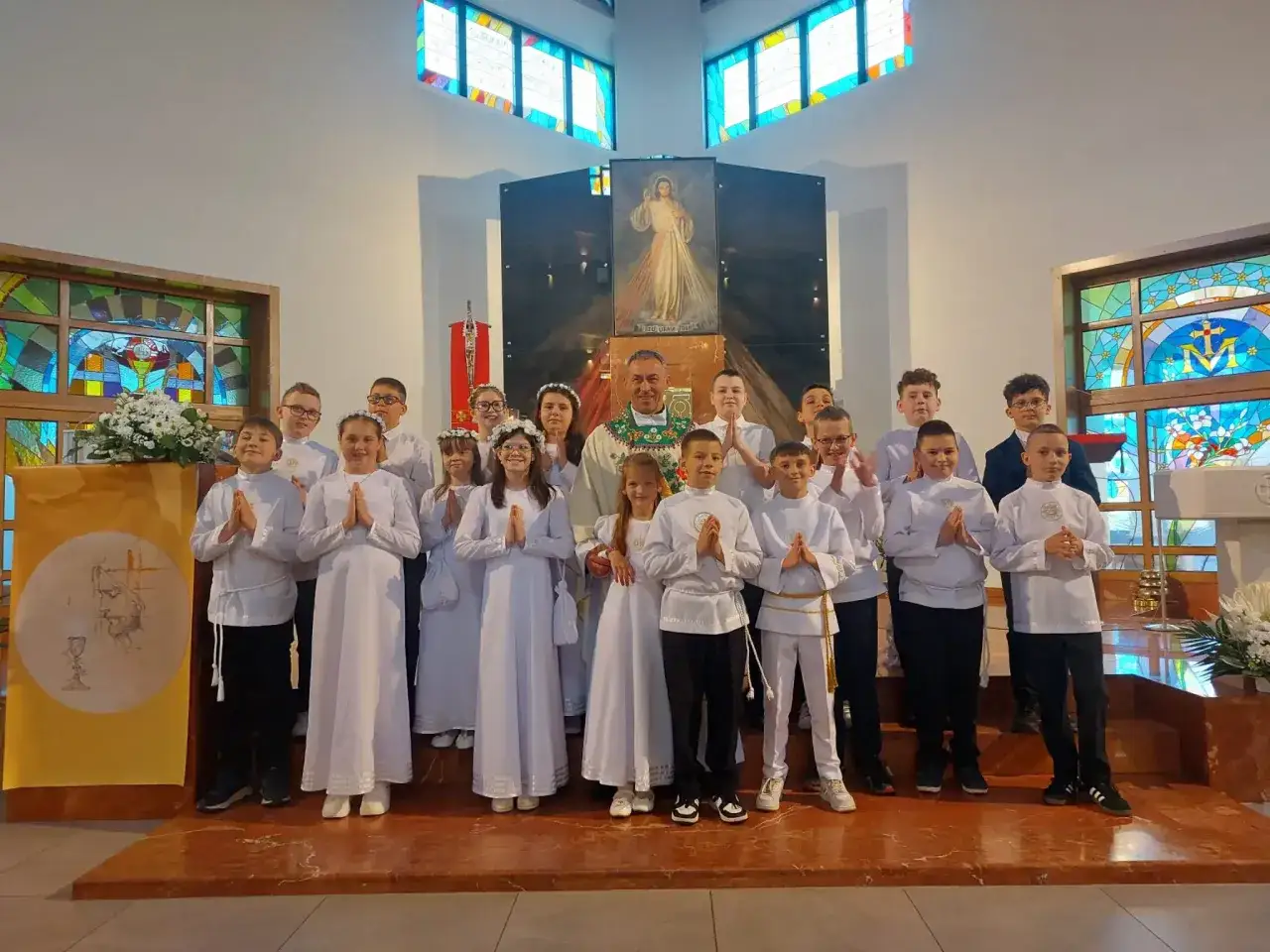 Dzieci w białych strojach komunijnych z księdzem w kościele. Czy rocznica komunii jest obowiązkowa? Ważne wydarzenie.