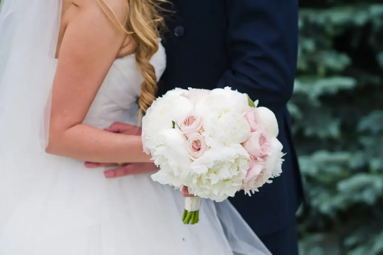 Piękny bukiet ślubny piwonie i eustoma w dłoniach pary młodej. Delikatne białe i różowe kwiaty tworzą romantyczną kompozycję.