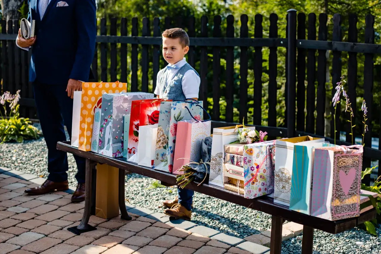 Chłopiec w eleganckim stroju stoi obok stołu pełnego prezent&oacute;w, pokazując, co zamiast kwiat&oacute;w na ślub może być świetnym pomysłem.