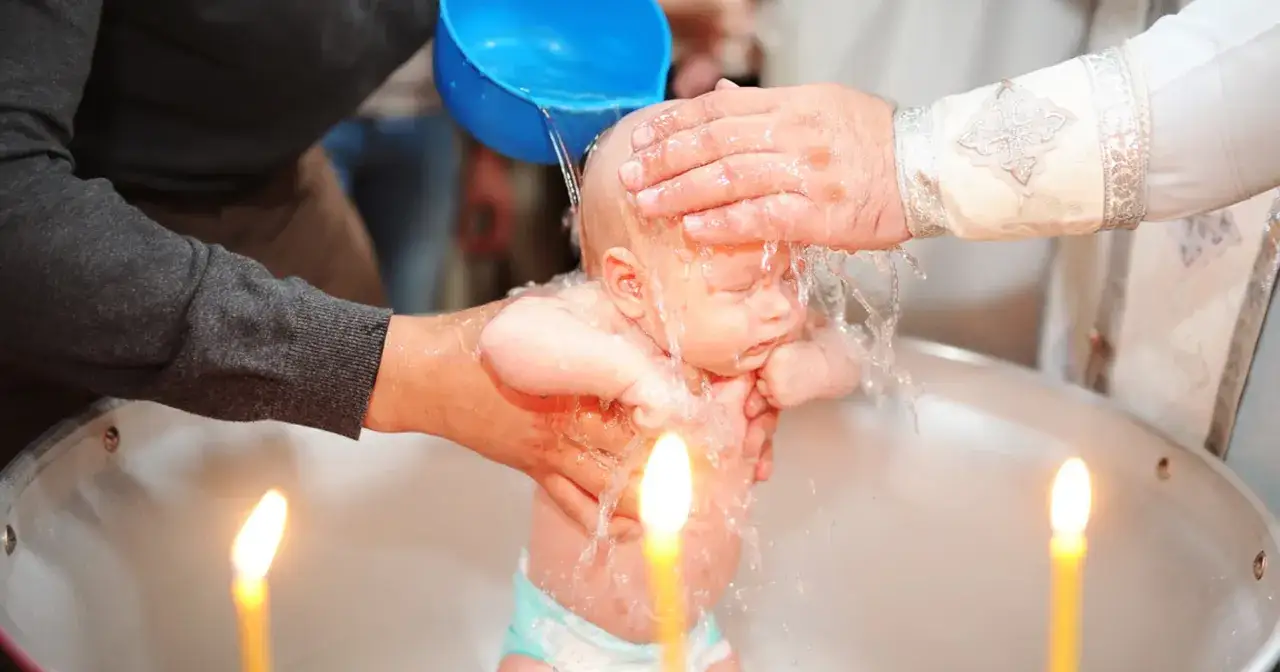 Chrzest krok po kroku: kapłan polewa wodą głowę niemowlęcia w chrzcielnicy, obok płoną świece.