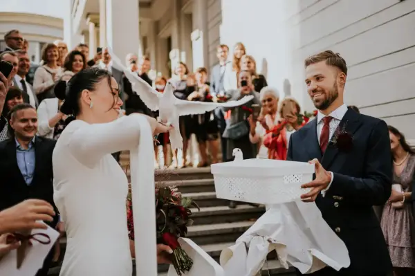 Para młoda wypuszcza gołębie na kameralnym weselu. W tle widać gości z telefonami.